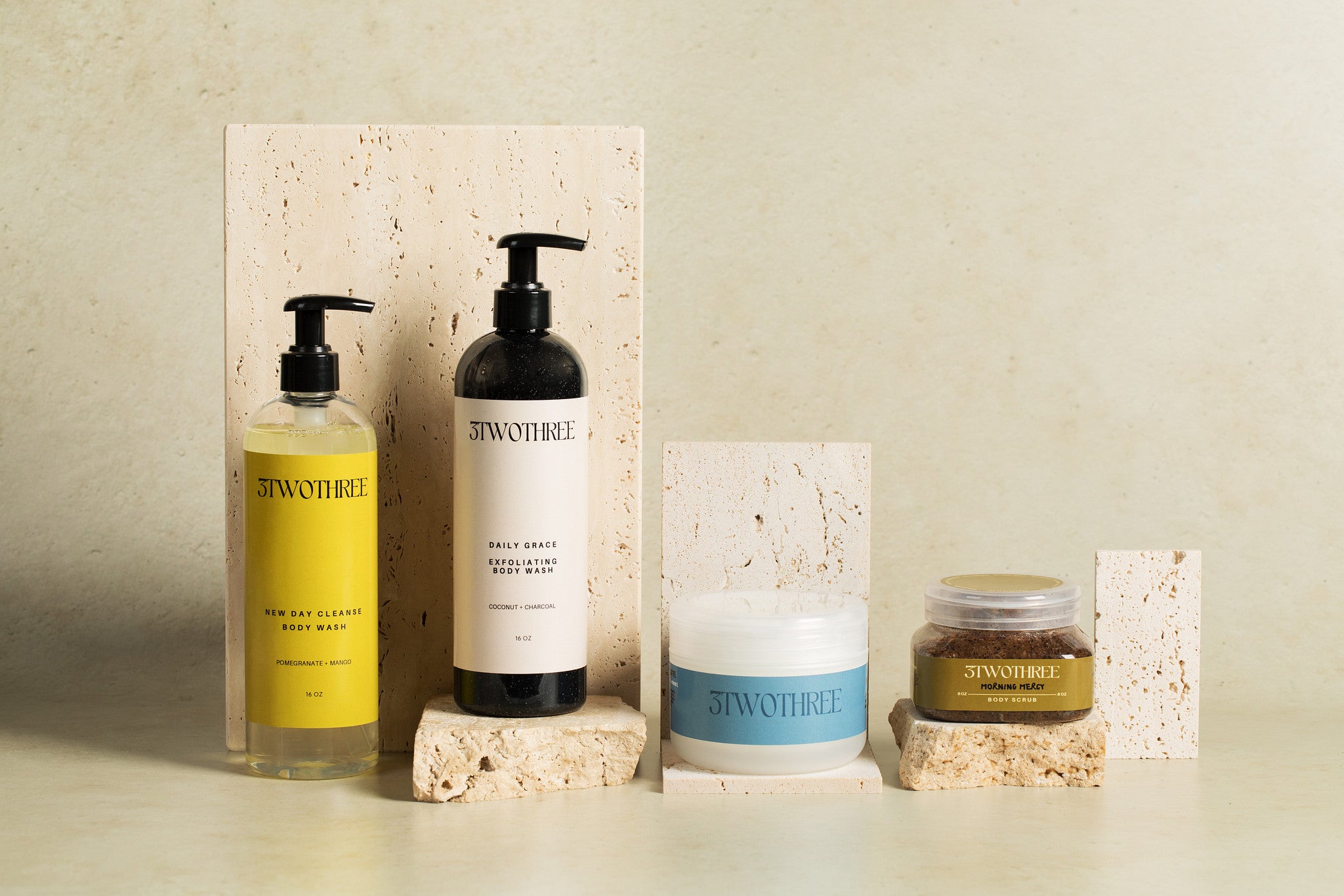 Set of '3TWOTHREE' body care products including a bottle of cleansing body wash, a bottle of exfoliating body wash, a jar of Body Butter, and a jar Body Scrub on a neutral background